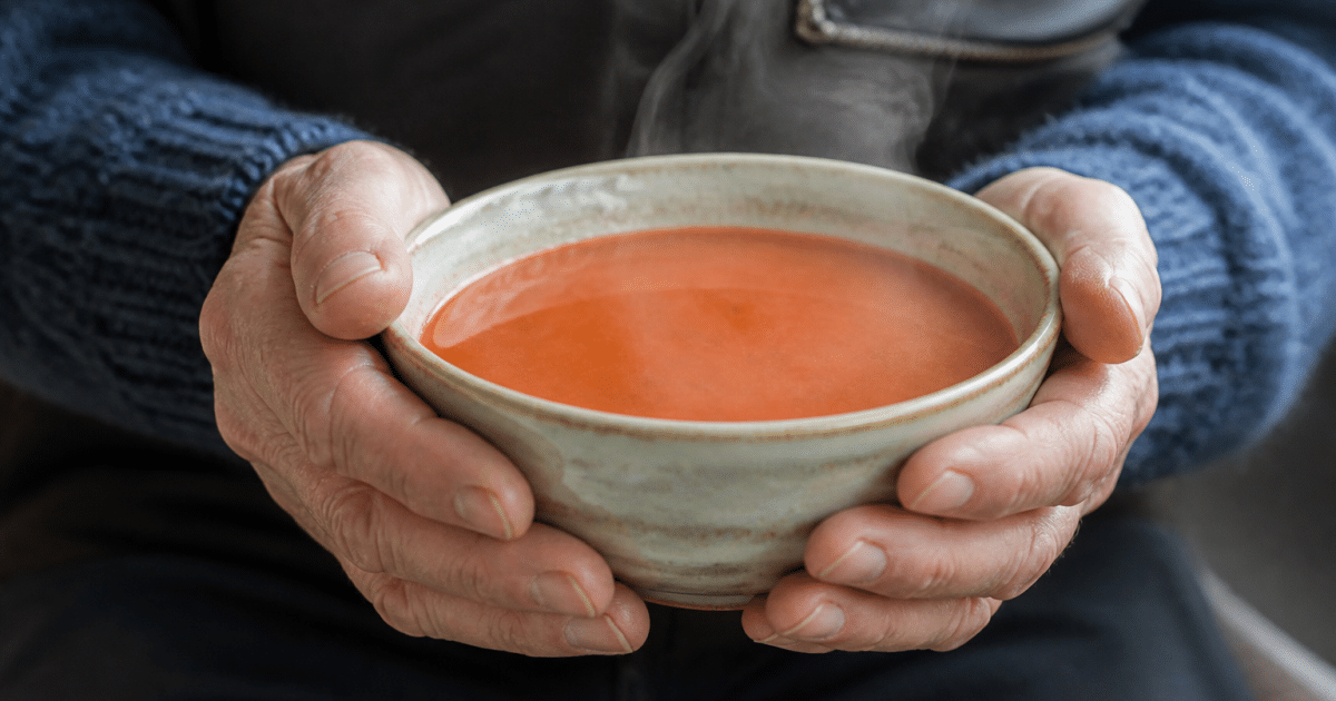 Bol de soupe tomate rouge fumante tenu par mains ridees